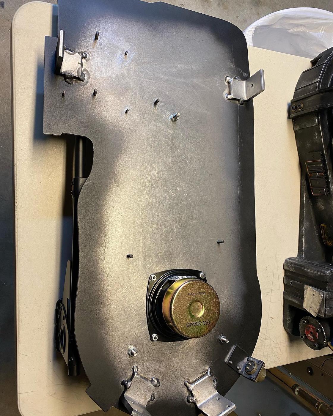 A fabricated steel panel with welded brackets and a mounted speaker on the workbench